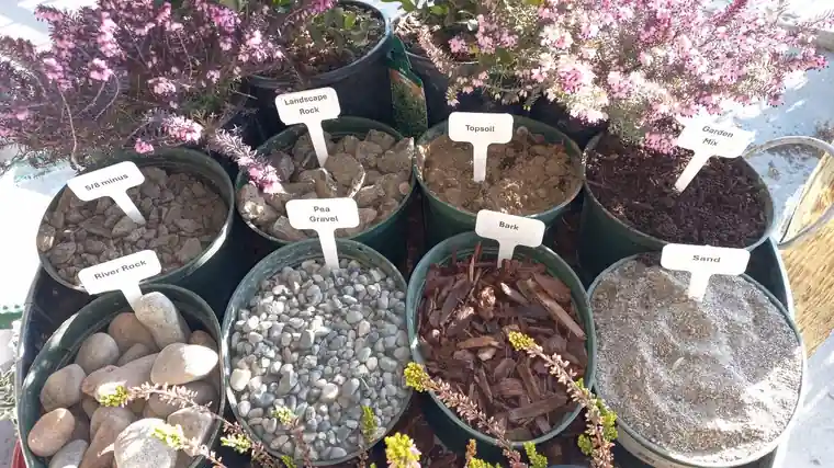 Sample tubs of decorative rock, river rock, bark and soil with handwritten labels at Roots