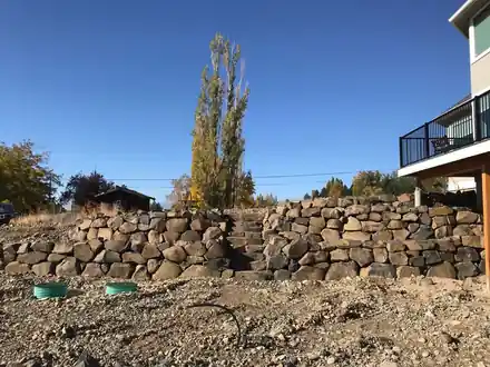 A stacked basalt rock wall under a house deck, bare ground in front of it