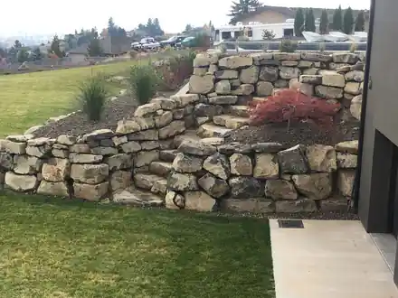 A stacked basalt boulder terrace with a red Japanese maple and ornamental grasses
