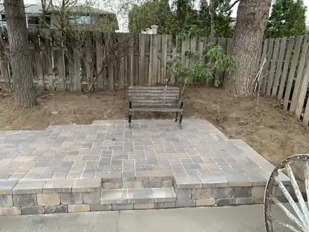 A raised paver patio with a bench against a board fence