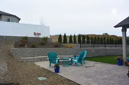 A terraced block-wall backyard with new shrub beds above a paver patio, fire pit and teal chairs