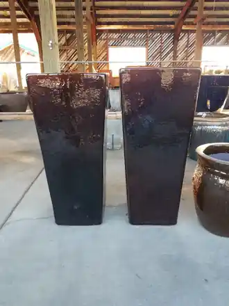 Two tall square dark glazed ceramic planters standing side by side on the concrete floor of the pottery barn
